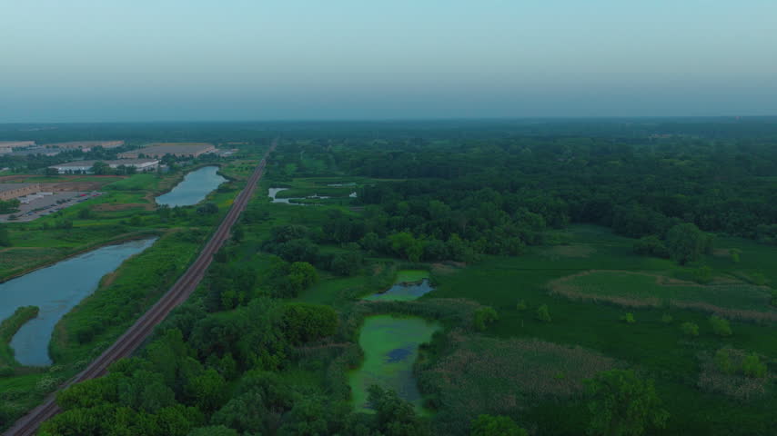Aerial view of train passing wetlands at dusk near Racine, Wisconsin. Drone shot along railroad tracks past marsh ponds, trees and an industrial park in summer blue hour light.