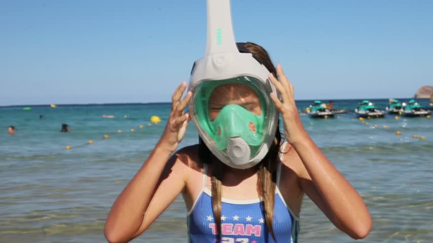 Young girl stands in shallow ocean water and adjusts a full face snorkeling mask, with swimmers, floating buoys, small boats, rocky coastline, and clear blue sky behind her