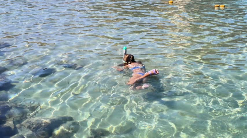 Young girl wearing a snorkel and fins swims steadily through transparent shallow seawater, with sunlight patterns, submerged rocks, sandy bottom, and floating boundary buoys visible