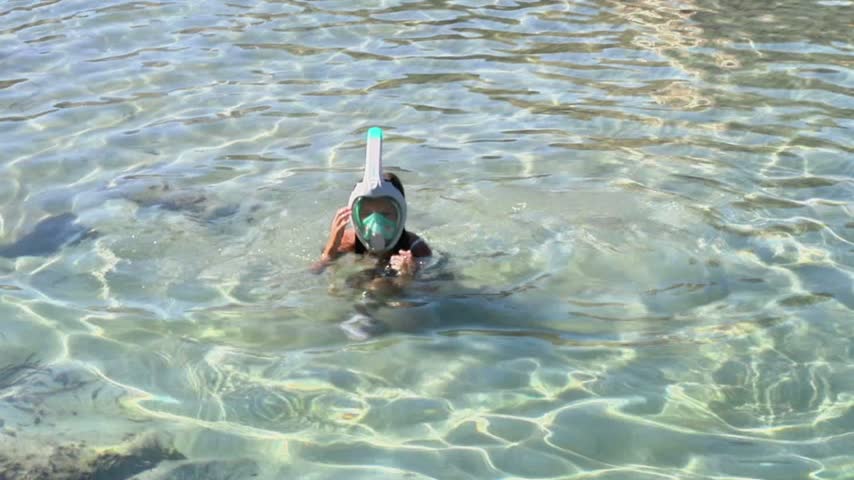 Young girl swimmer wearing a snorkeling mask and tube moves slowly through clear shallow seawater, with sunlight creating bright patterns across the surface and rocky seabed visible below