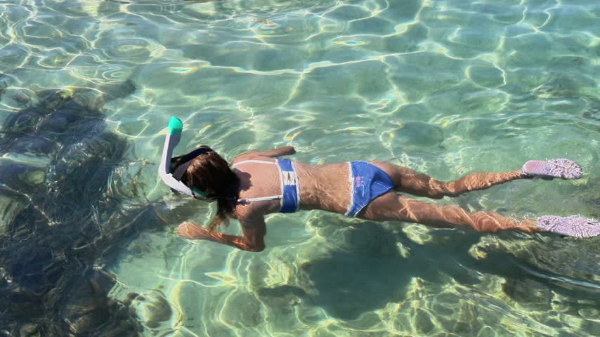 Young girl wearing blue swimwear and pink fins swims forward while snorkeling through clear shallow seawater, revealing rippling sunlight, submerged rocks, and textured seabed below