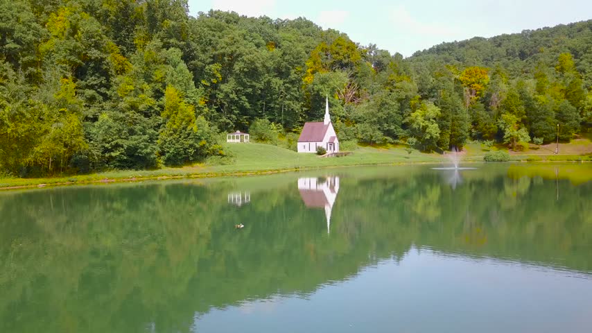 Small white church reflecting in calm lake water. Peaceful woodland landscape with chapel and fountain in morning light.