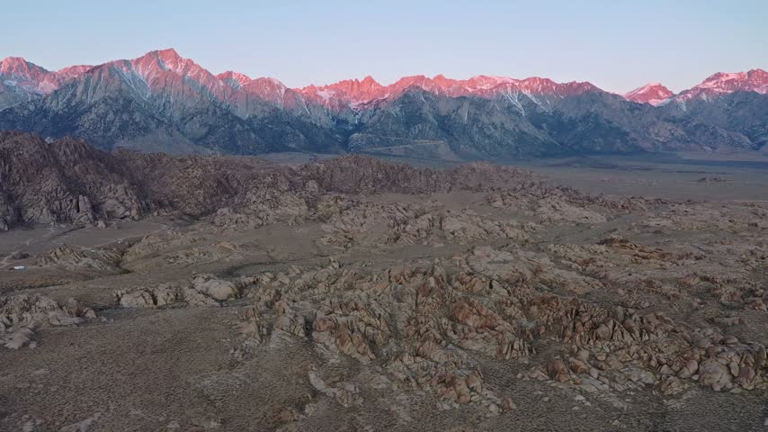 Aerial drone footage flying over rugged desert rocks of Alabama Hills with illuminated snowy Sierra Nevada mountain range at sunrise.