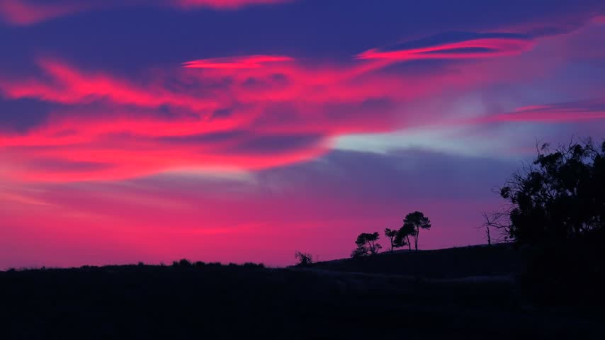 Dramatic sky with vibrant pink and purple lenticular clouds during dusk. Silhouetted landscape with trees and grass.