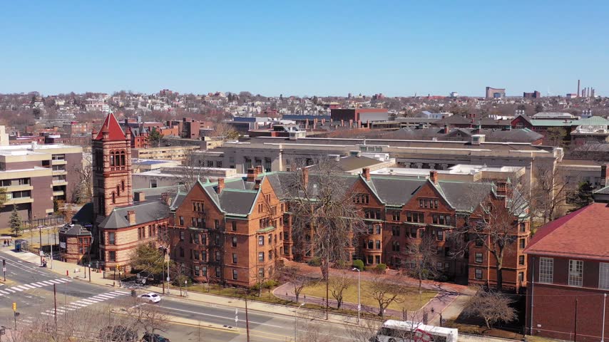 Slow drone panning shot of red brick collegiate architecture on bright sunny day with city skyline in background.