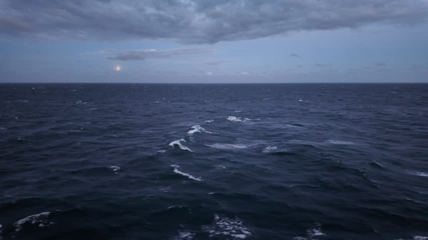 Looking out from a moving ship towards the horizon at night. The moon is low in the sky, and the sea conditions are calm. There are clouds in the sky above.