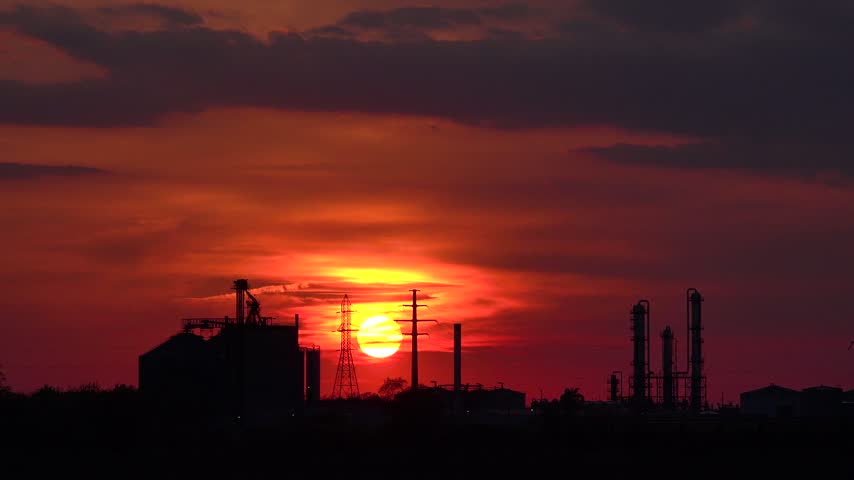 Stunning sunset over industrial factory silhouettes with dramatic red clouds and glowing sun setting behind power poles.