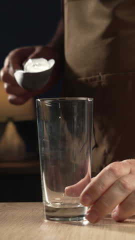 Man in apron adding ice cube to glass on wooden table, creating a refreshing beverage. Close-up