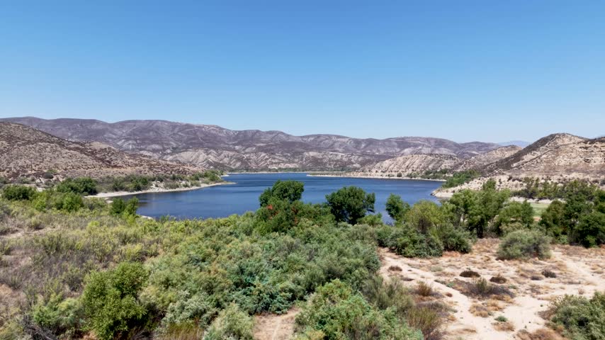 Vail Lake framed by dense shrubs and trees against dry rugged hills beneath a clear blue sky, aerial establishing dolly flyover
