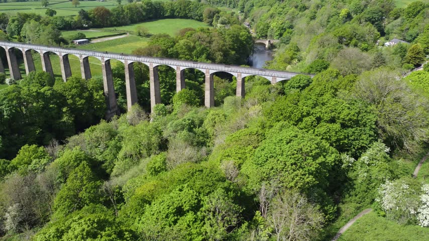 Aerial drone flight over dense spring woodland towards Pontcysyllte Aqueduct, the longest aqueduct in Britain, carrying the Llangollen Canal on tall stone piers and iron arches above the River Dee near Wrexham in North Wales. The camera passes over the cast iron trough and towpath to look down on the river winding through the wooded gorge, with an old stone road bridge and green fields beyond in bright sunshine.