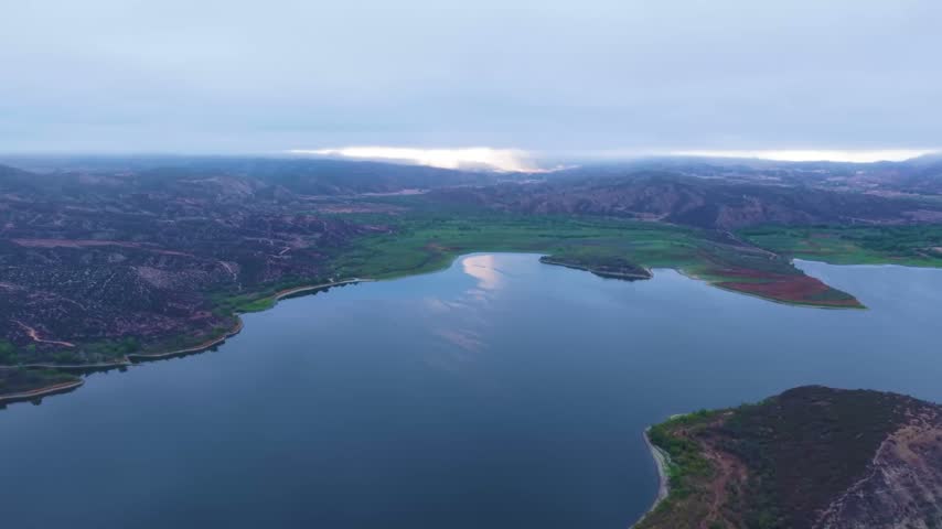 Vail Lake spreads between rugged hills beneath a low cloud layer and muted overcast sky, aerial panoramic ascend into misty clouds