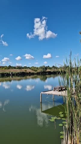 Picturesque lake under blue sky with clouds, place for fishing, country vacation, unity with nature. Landscape with reflection of clouds in water, reeds and village houses on the far shore