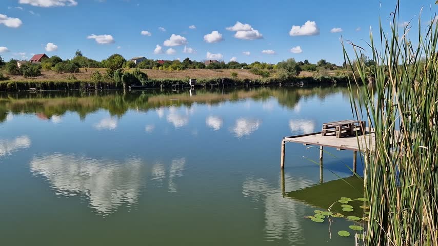 Picturesque lake under blue sky with clouds, place for fishing, country vacation, unity with nature. Landscape with reflection of clouds in water, reeds and village houses on the far shore