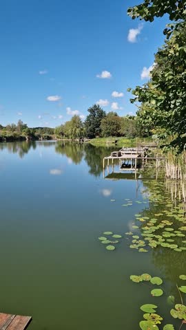 Picturesque lake under blue sky with clouds, place for fishing, country vacation, unity with nature. Landscape with reflection of clouds in water, reeds and village houses on the far shore