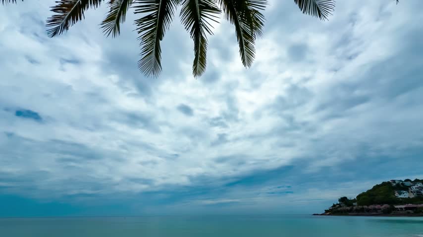 A view over the ocean with clouds changing above the water. Palm trees can be seen framing the scene along the coast.