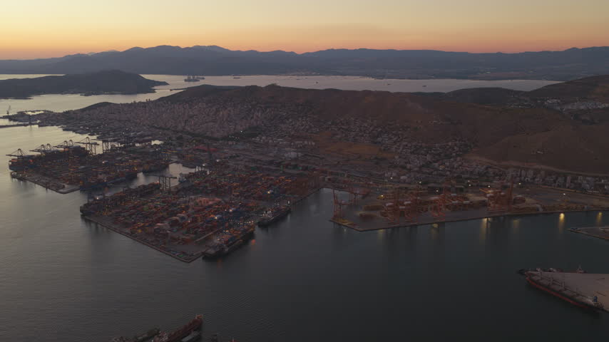 Expansive aerial footage showcasing the Port of Piraeus, Greece, with its bustling container terminal, cargo ships, and dockside cranes illuminated against a beautiful sunset sky