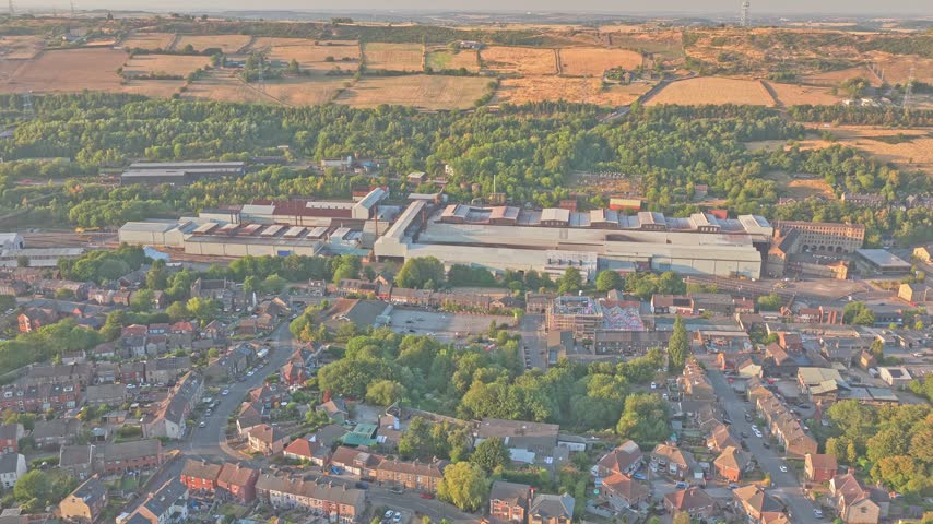 Stocksbridge steelworks dominates valley beside dense housing, woodland and cultivated upland fields, South Yorkshire, England, with extensive production halls, drone forward flyover.