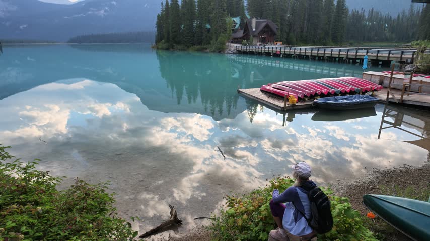 top view of woman by turquoise waters of Emerald Lake,admiring reflections of mountains and clouds,with boat dock and red canoes, enjoying summer travel in Yoho National Park, British Columbia, Canada