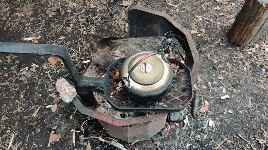 A traditional metal kettle boiling water over a warm campfire inside a sturdy metal fireplace setup on the natural forest floor in the wilderness of Lapland, Finland, showcasing outdoor cooking and peaceful ecology of the northern region