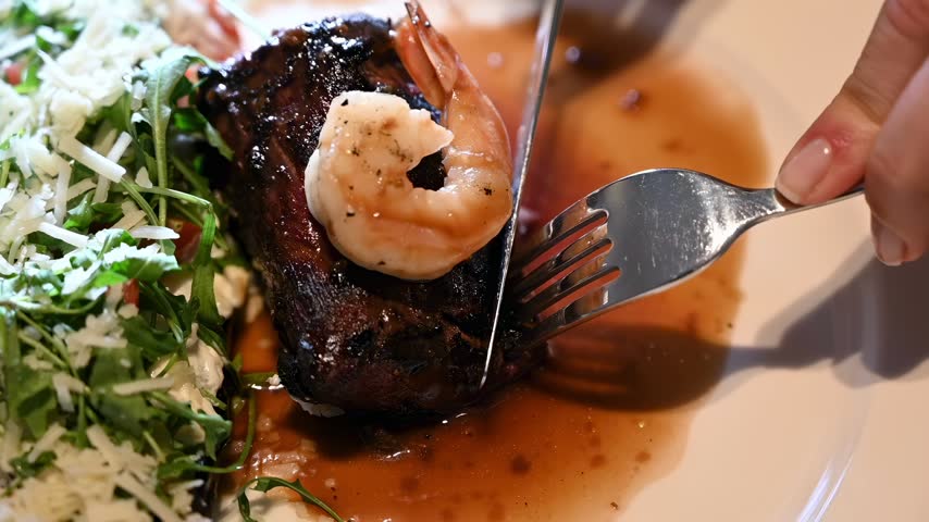 Close up shot showing person slicing juicy grilled steak topped with shrimp served with salad and gourmet sauce on white plate.