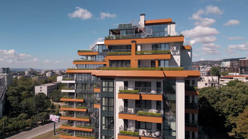 Exterior of a modern multi-story residential building with orange facade elements, green terraces, penthouses, and large glass windows under a sunny blue sky.