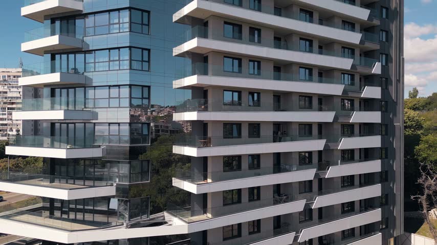 Close-up exterior view of a high-rise modern residential building with white balconies, glass facade reflecting the cityscape, contemporary architecture design.