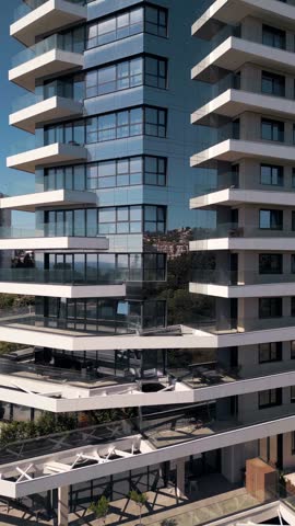 Close-up exterior view of a high-rise modern residential building with white balconies, glass facade reflecting the cityscape, contemporary architecture design.