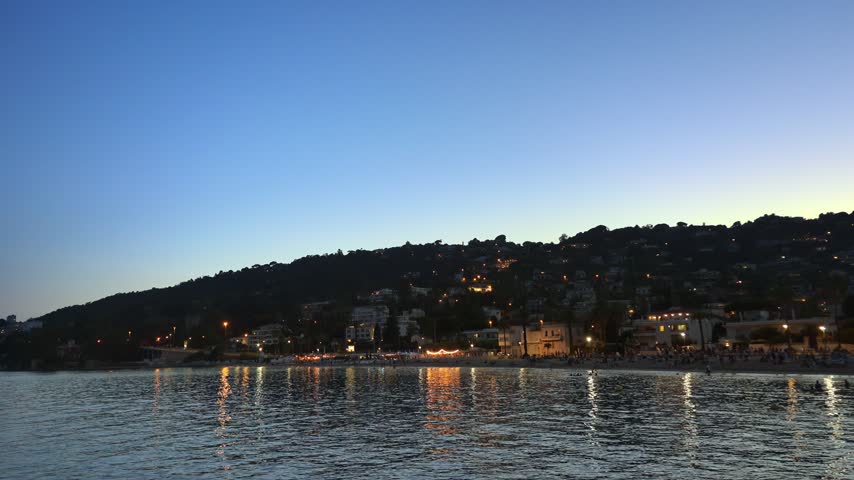 Breathtaking dusk view of seaside town nestled on hillside featuring twinkling city lights reflecting over calm ocean water.