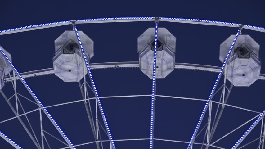 Close up of illuminated white ferris wheel at night, spinning amusement park ride, fairground attraction with blue sky.