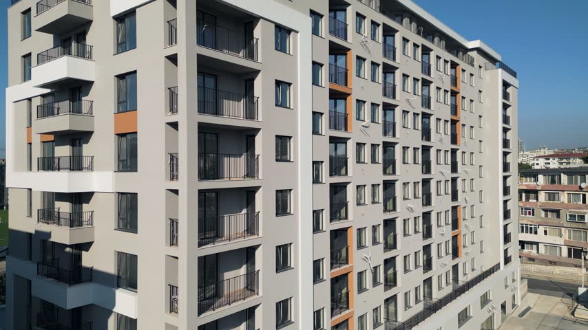 Exterior view of a modern multi-story residential apartment building with grey and orange facade design, balconies, and windows under clear blue sky. Contemporary urban housing complex.