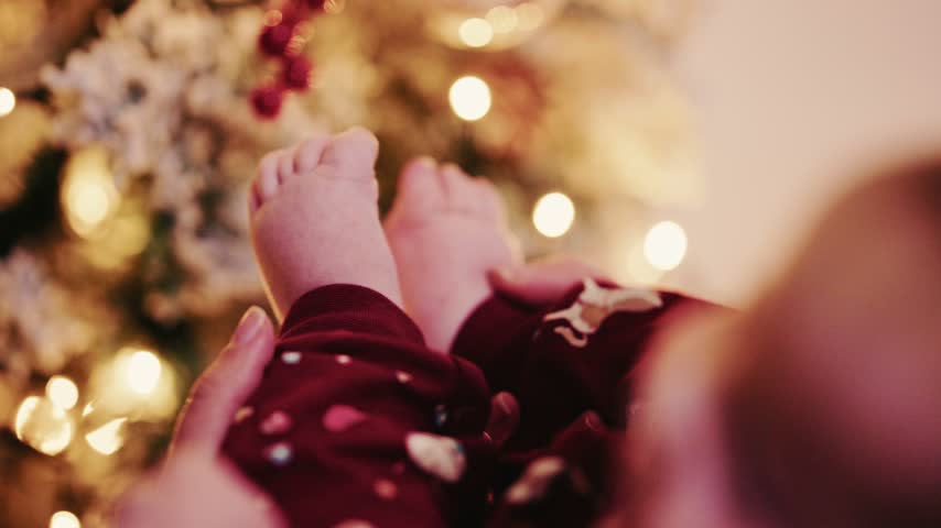 Close up of infant legs moving in red pajamas against bokeh background of decorated pine tree and warm winter lights.