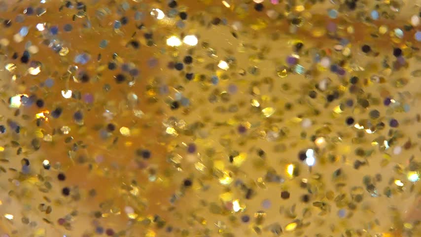 A vibrant macro view of orange and golden glitter sparkling under warm light, with reflective particles producing colorful highlights and a soft shimmering effect.