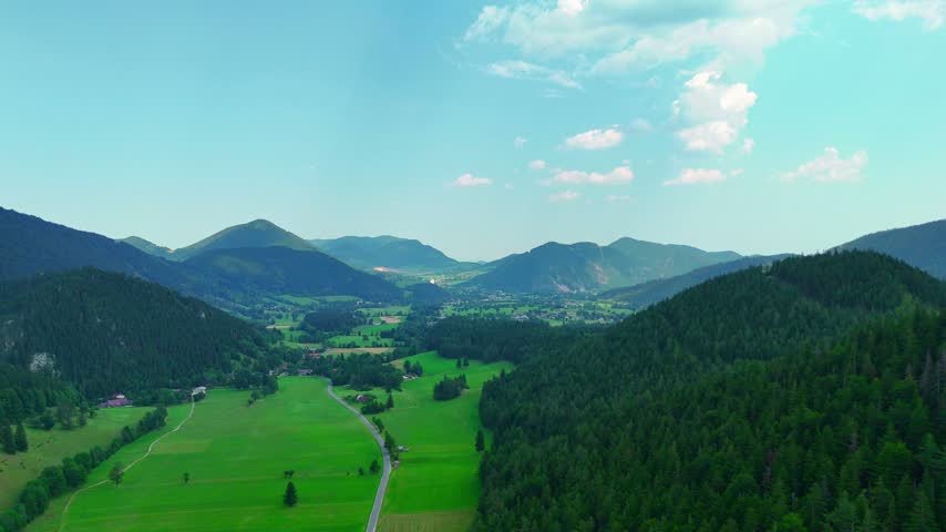 Mountain landscape with green fields and hills under a blue sky. Hills and green fields with some trees and a road. The sky is bright and blue.