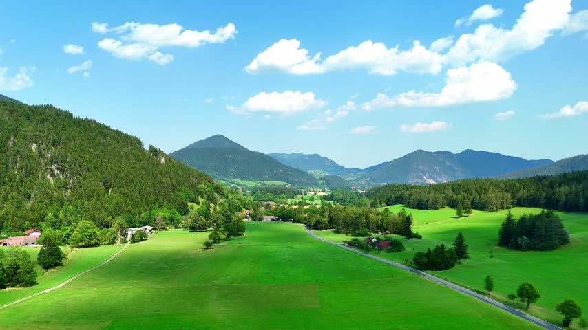 Green fields and mountains under a blue sky in a rural area. Wide green fields with trees and mountains. The sky is clear with clouds.