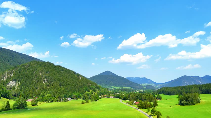 Green landscape with mountains under a blue sky on a sunny day. Mountains surround a green valley with trees and small houses visible in the distance.