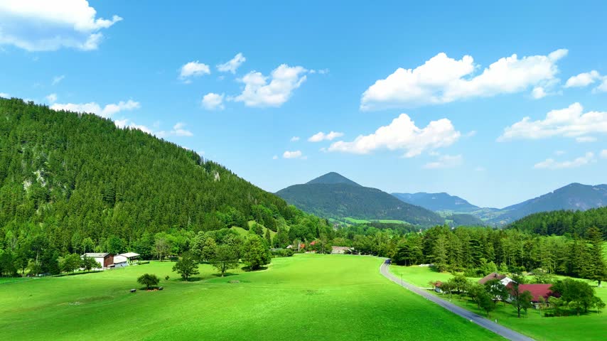 Mountain landscape with green fields and clear blue sky in the countryside. Green fields stretch below mountains under a blue sky with some clouds. Trees dot the area.