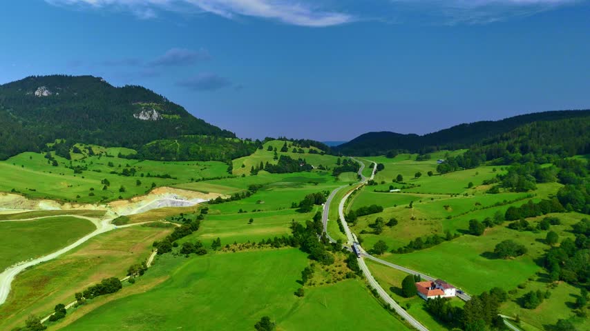 Views of rolling green hills and winding road in a landscape with mountains. Green hills stretch across the scene with a winding road and a house nearby. The sky is clear.