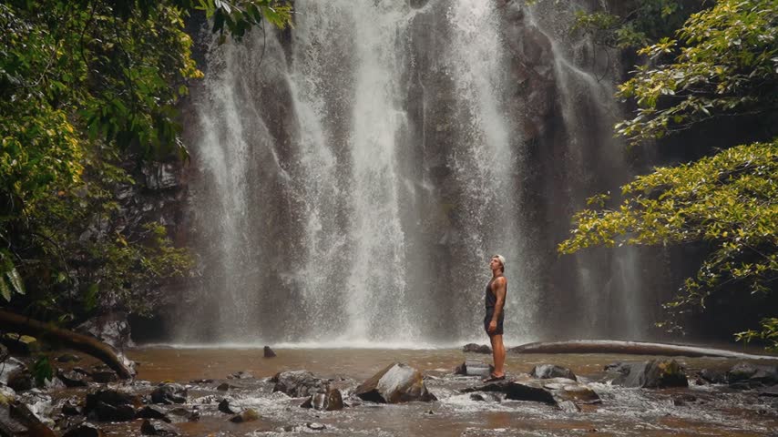 Cinematic footage of a young man standing in front of a huge waterfall in a rainforest.