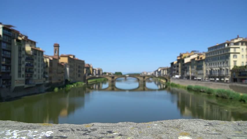 Wide view of Ponte Vecchio spanning Arno River with historic buildings, calm water, stone bridge arches and clear blue sky creating scenic landscape across Florence Italy
