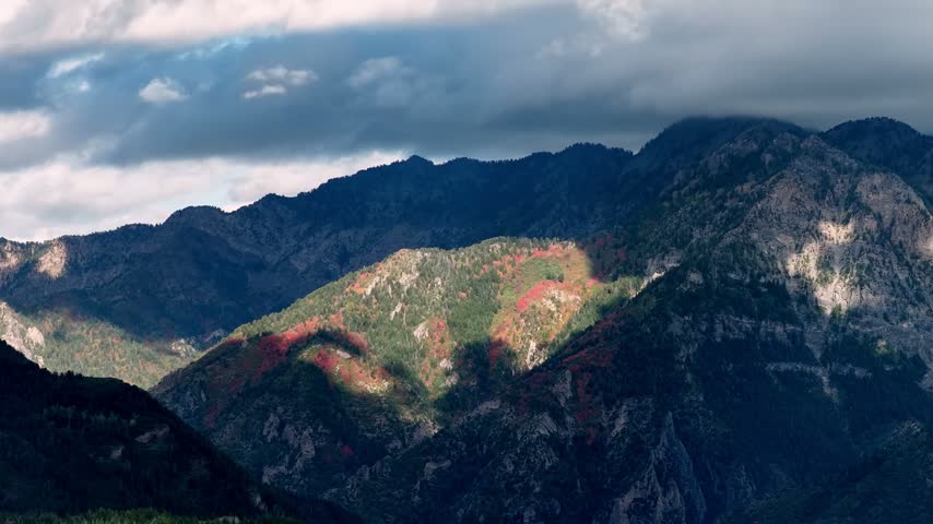 Clouds moving over a rugged alpine forest with vivid autumn colors and shifting shadows dancing across the steep mountainous terrain in a static time lapse.