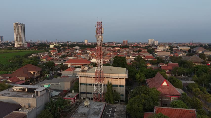 SURABAYA, INDONESIA - SEPTEMBER 23, 2026: Aerial orbit around a red and white telecommunications tower above a dense residential neighborhood with distant high-rise apartments