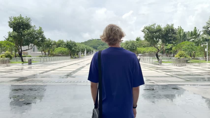 Young Blond Man Walking on Wide Park Path in Shenzhen and Talking on Smartphone – Back View with Tropical Greenery and Blue Sky	
