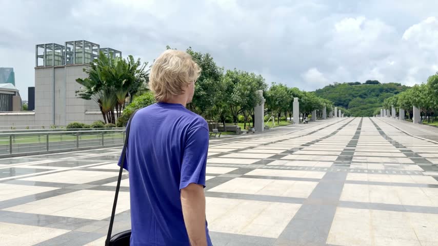 Young Blond Man Walking on Wide Park Path in Shenzhen and Talking on Smartphone – Back View with Tropical Greenery and Blue Sky	
