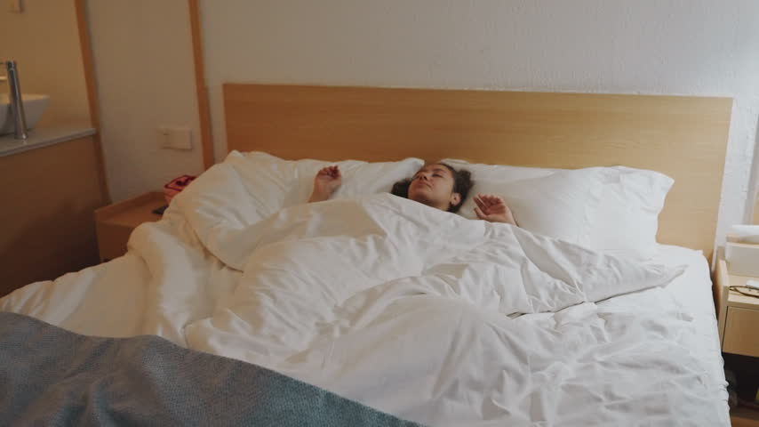 Bleary female guest in hotel bed, head on pillows under white duvet surrounded by wood headboard and sink corner slow waking sequence shows hand reaching to nightstand, restless shifting, soft morning
