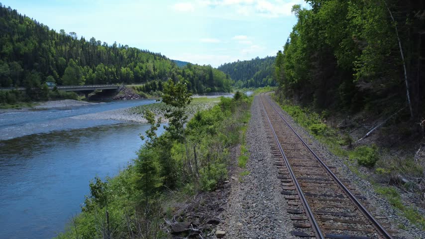 Sweeping aerial imagery follows a railway line bordering the Matapedia River while a concrete bridge stands prominently within the surrounding forest landscape.