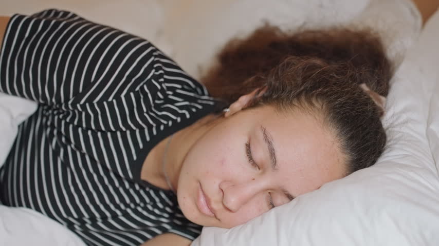 Hotel room afternoon young woman napping on crisp white sheets in striped shirt, soft natural light filters through curtains, guest stretches arm and adjusts pillow while smartwatch and bracelet peek,