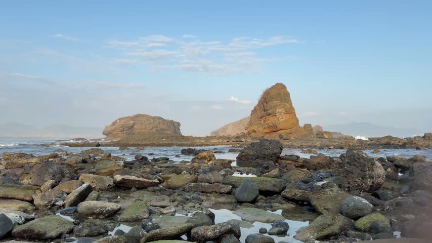 A serene coastal landscape featuring large rock formations and scattered boulders along the shore under a clear blue sky.
