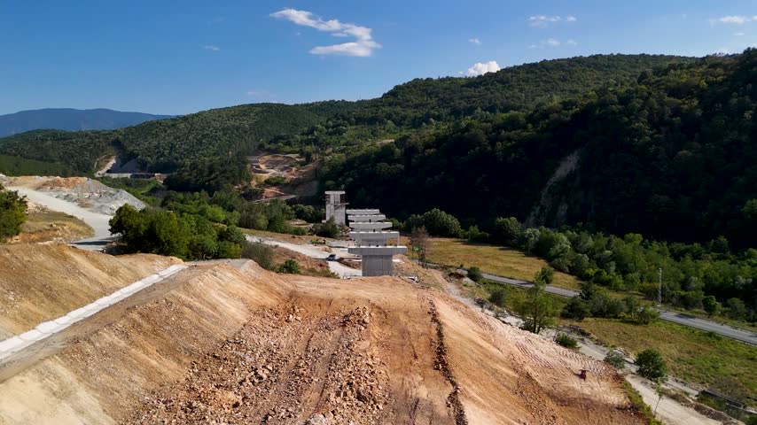 Aerial view of unfinished concrete bridge pillars crossing a rural mountain valley, surrounded by forested hills, winding roads and clear blue sky
