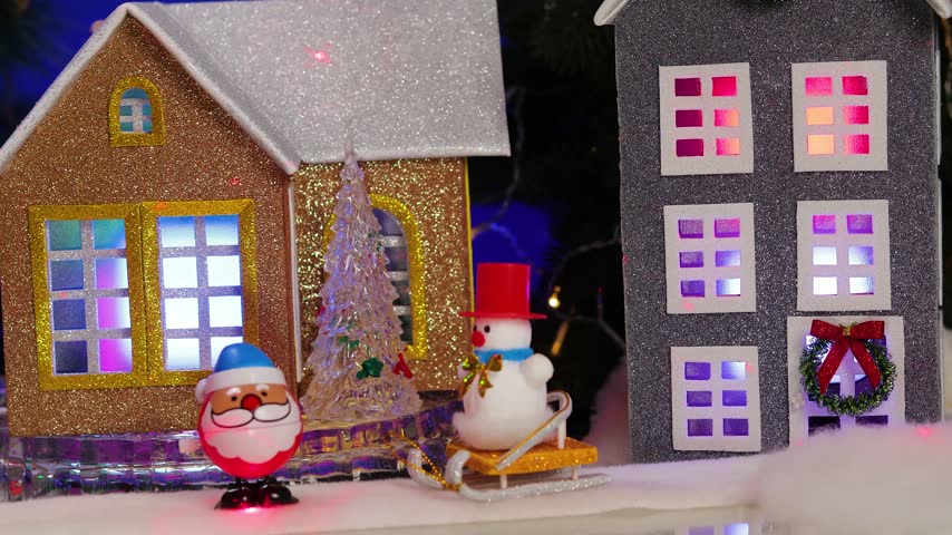 In the center of the Christmas village, a yellow car pulls up to a gray little house adorned with small fir trees and snowmen