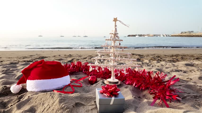 Christmas decorations, including a Santa hat, a small tree, and a gift box, rest on the sand of a tropical beach, with the calm ocean and sailboats visible in the background at sunrise
